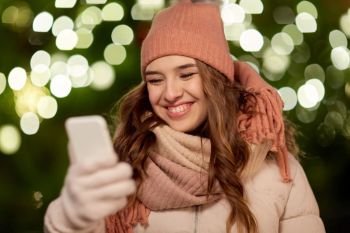 holidays, communication and people concept - portrait of beautiful happy young woman with smartphone over christmas lights in winter evening. happy woman with smartphone at christmas