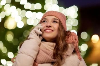 holidays, communication and people concept - portrait of beautiful happy young woman calling on smartphone over christmas tree lights in winter evening. happy woman calling on smartphone at christmas