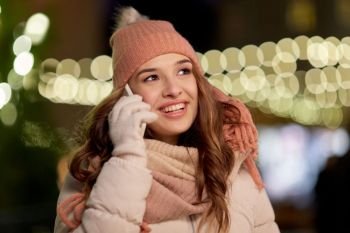 holidays, communication and people concept - portrait of beautiful happy young woman calling on smartphone over christmas lights in winter evening. happy woman calling on smartphone at christmas