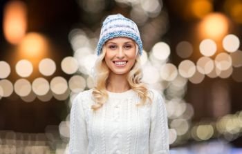 holidays, christmas and people concept - smiling young woman in winter hat, sweater and gloves over night lights background. happy woman over christmas lights