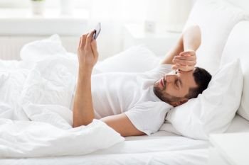 technology, internet, communication and people concept - happy young man texting on smartphone in bed at home in morning. young man with smartphone in bed in morning