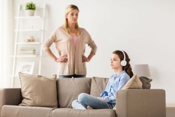 people, problem and family concept - girl with earphones sitting on sofa and angry mother at home. girl with earphones and angry mother at home