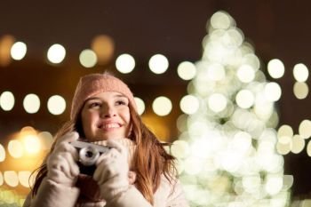 holidays and people concept - beautiful happy young woman with camera over christmas tree lights in winter evening. happy young woman with camera over christmas tree