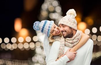 christmas, holidays and people concept - happy couple in hats and scarf hugging over night lights background. happy couple hugging over christmas lights