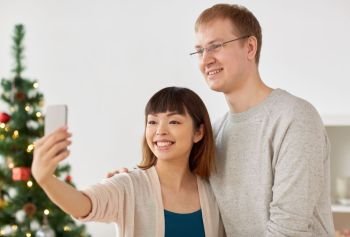 winter holidays, technology and people concept - happy mixed-raced couple taking selfie by smartphone home at christmas. mixed-raced couple taking selfie at christmas