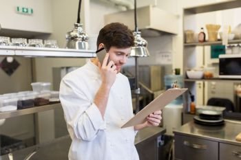 cooking, profession and people concept - male chef cook with clipboard calling on smartphone at restaurant kitchen. chef cook calling on smartphone at restaurant kitchen