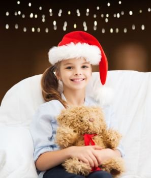 christmas, holidays and people concept - happy girl in santa hat with teddy bear over garland lights background. girl in santa hat with teddy bear at christmas