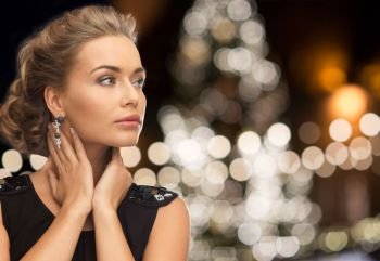 jewelry, holidays, luxury and people concept - beautiful woman in black wearing earrings over christmas tree lights background. woman wearing jewelry over christmas lights