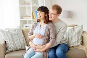 pregnancy, family and people concept - happy pregnant wife with husband sitting on sofa at home. happy pregnant wife with husband at home