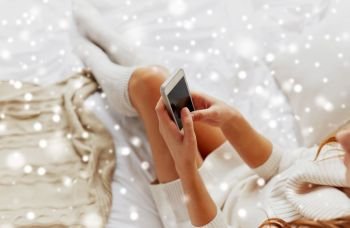 technology, communication, winter and people concept - happy young woman lying in bed and texting on smartphone at home bedroom over snow. happy young woman with smartphone in bed at home