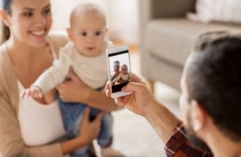 family, parenthood and people concept - happy father with smartphone taking picture of mother with baby at home. happy family with baby photographing at home
