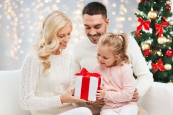 family, christmas, holidays and people concept - happy mother, father and little daughter with gift box sitting on sofa over lights background. happy family at home with christmas gift box