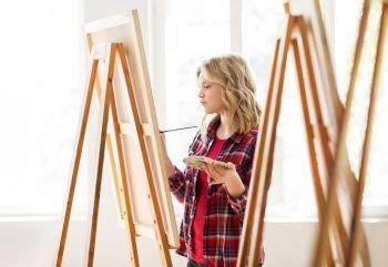art school, creativity and people concept - student girl or artist with easel, palette and paint brush painting at studio. student girl with easel painting at art school