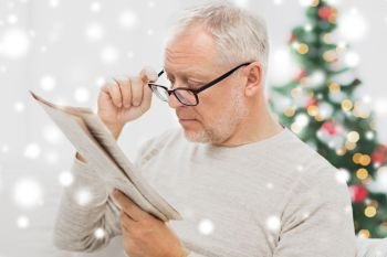 holidays, information, people, vision and mass media concept - senior man in glasses reading newspaper at home over christmas tree background and snow. senior man in glasses reading newspaper at home
