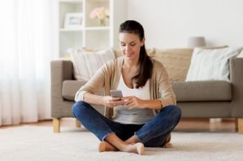 people, technology, communication and leisure concept - happy young woman sitting on floor and texting message on smartphone at home. happy woman texting message on smartphone at home
