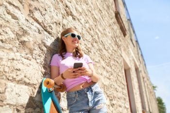 lifestyle, summer, technology and people concept - smiling young woman or teenage girl in sunglasses with longboard and smartphone over stone wall outdoors. happy teenage girl with longboard and smartphone