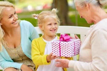 family, holidays and people concept - happy smiling granddaughter giving present to her grandmother sitting on park bench. happy family giving present to grandmother at park
