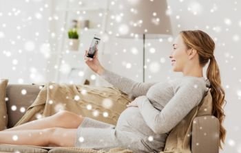 pregnancy, motherhood, technology, people and expectation concept - happy pregnant woman with smartphone taking selfie at home over snow. pregnant woman taking smartphone selfie at home
