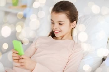 people, children and technology concept - happy smiling girl lying awake with smartphone in bed over holidays lights background. happy girl in bed with smartphone over lights