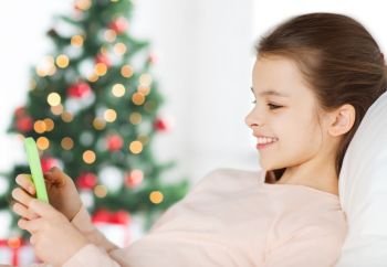 holidays, children and technology concept - happy smiling girl with smartphone lying in bed at home over christmas tree background. happy girl in bed with smartphone at christmas
