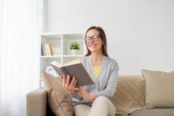 literature, education and people concept - happy middle-aged woman in glasses reading book at home. happy woman in glasses reading book at home