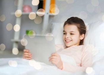 people, children and technology concept - happy smiling girl lying awake with tablet pc computer in bed at home over holidays lights. happy girl lying in bed with tablet pc at home