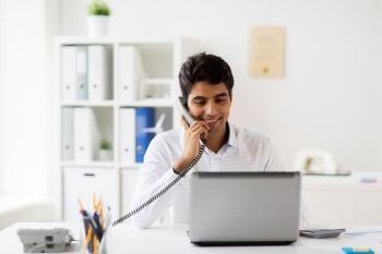 business, communication and people concept - happy businessman with laptop computer calling on desk phone at office. happy businessman calling on desk phone at office