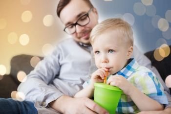 family, childhood, fatherhood, care and people concept - father helping little son with drinking from cup at home. father and son drinking from cup at home