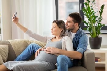 technology, pregnancy and people concept - happy man and his pregnant wife taking selfie by smartphone at home. man and pregnant woman taking selfie at home