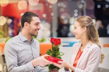 love, romance, valentines day, couple and people concept - happy young man with red flowers giving present to smiling woman at cafe in mall. happy couple with present and flowers in mall