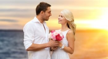 love, wedding and people concept - happy couple in white clothes with flowers over sea background. happy couple with flowers over sea background