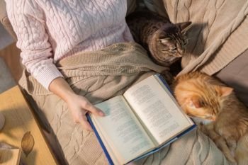 hygge, literature and people concept - close up of red and tabby cat and female owner reading book in bed at home. red and tabby and owner reading book at home