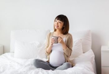 pregnancy, rest, people and expectation concept - happy pregnant asian woman with cup drinking tea in bed at home bedroom. happy pregnant woman with cup drinking tea at home
