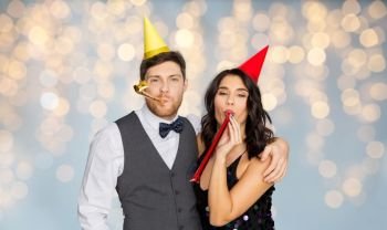 birthday, celebration and holidays concept - happy couple with party blowers and caps having fun over festive lights background. happy couple with party blowers having fun