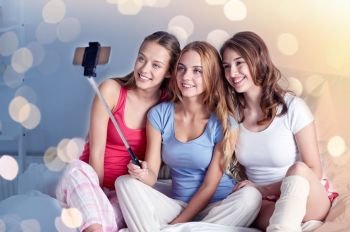 friendship, people, pajama party and technology concept - happy friends or teenage girls with smartphone and monopod taking selfie at home. teen girls with smartphone taking selfie at home