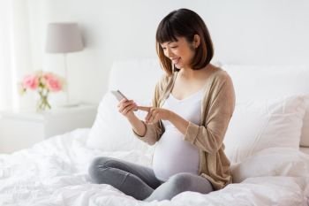 pregnancy, technology and people concept - happy pregnant asian woman with smartphone in bed at home. pregnant woman with smartphone in bed at home