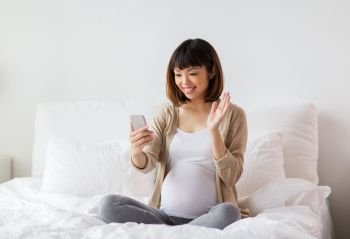 pregnancy, technology and people concept - happy pregnant asian woman taking selfie by smartphone in bed at home and waving hand. pregnant woman taking selfie by smartphone at home