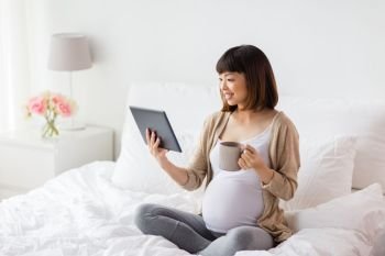 pregnancy, technology, people and expectation concept - happy pregnant asian woman with tablet pc computer drinking tea in bed at home. happy pregnant asian woman with tablet pc at home