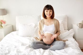 pregnancy, people and health care concept - happy pregnant asian woman sitting in bed at home with pills and glass of water. happy pregnant woman with pills in bed at home