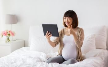 pregnancy, technology and people concept - happy pregnant asian woman with tablet pc computer having video call and showing thumbs up in bed at home. happy pregnant asian woman with tablet pc at home
