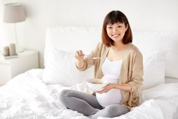 pregnancy, people and breakfast concept - happy pregnant asian woman eating cereal flakes in bed at home bedroom. happy pregnant woman eating cereal flakes at home