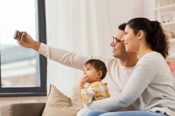 family, technology, parenthood and people concept - happy mother and father with baby daughter taking selfie by smartphone at home. mother and father with baby taking selfie at home