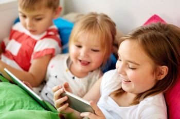 childhood, technology and family concept - happy little kids with smartphone and tablet pc computer in bed at home. little kids with smartphone in bed at home