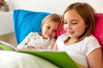 childhood, leisure and family concept - little girls or sisters reading book in bed. little girls or sisters reading book in bed