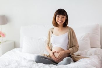 pregnancy, love, people and expectation concept - happy pregnant asian woman sitting in bed and making hand heart gesture at home. happy pregnant woman making heart gesture in bed