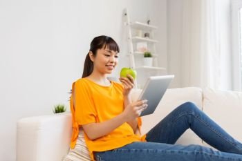 technology, education, diet and people concept - happy young asian woman sitting on sofa with tablet pc computer and green apple at home. happy asian woman with tablet pc and apple at home