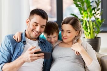 family, pregnancy and technology concept - happy pregnant mother, father and little son with smartphone sitting on on sofa at home. happy family with smartphone at home