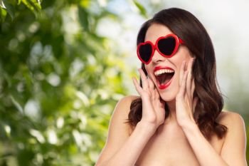 valentines day, beauty and people concept - happy smiling young woman with red lipstick and heart shaped sunglasses over green natural background. woman with red lipstick and heart shaped shades