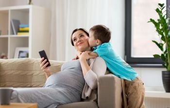 family, emotions and pregnancy concept - little son kissing happy pregnant mother with smartphone at home. son kissing happy pregnant mother at home