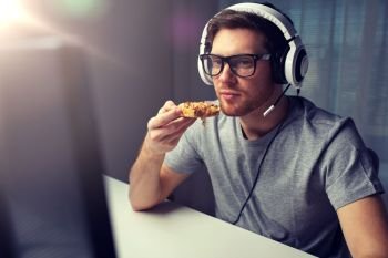 technology, gaming, entertainment, let’s play and people concept - young man in headset with pc computer eating pizza while playing game at home and streaming playthrough or walkthrough video. man in headset playing computer video game at home